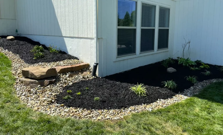 A freshly landscaped garden bed around the foundation of a home featuring dark mulch, green plants, and stones.